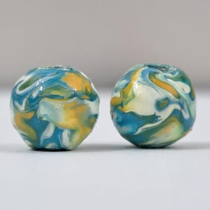 May include: Two round beads with a marbled design. The beads feature swirls of blue, yellow, white, and green. The beads are likely for jewelry making or crafts.
