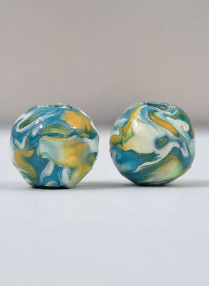 May include: Two round beads with a marbled design. The beads feature swirls of blue, yellow, white, and green. The beads are likely for jewelry making or crafts.