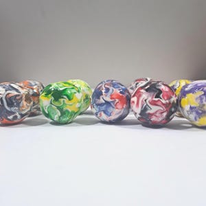 Puede incluir: Una colección de esferas de mármol coloridas. Las esferas muestran una variedad de colores, incluyendo rojo, azul, verde, amarillo, negro y blanco, arremolinados juntos en un patrón decorativo. Las esferas están dispuestas en una fila.