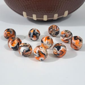 May include: Round beads with a marbled pattern in orange, black, and white. The beads are scattered on a white surface, with a brown football-shaped object in the background.