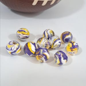 May include: Round beads with a marbled pattern in purple, yellow, and white. A brown American football with white stitching is in the background. The beads are on a white surface.