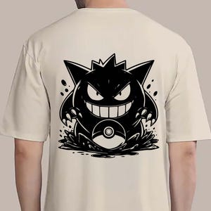 May include: A cream-colored t-shirt featuring a black graphic of a cartoon monster with sharp teeth and claws. The monster is positioned above a stylized splash, with a pokeball visible. The design is centered on the back of the shirt.