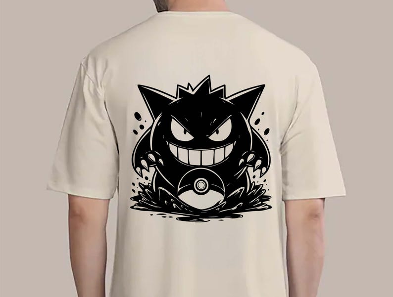 May include: A cream-colored t-shirt featuring a black graphic of a cartoon monster with sharp teeth and claws. The monster is positioned above a stylized splash, with a pokeball visible. The design is centered on the back of the shirt.