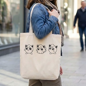 May include: Cream-colored tote bag featuring three black line art illustrations of a cartoon character with large eyes and a swirl on its head. The bag has a simple design with long handles for carrying over the shoulder.
