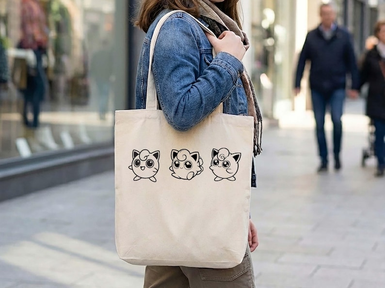 May include: Cream-colored tote bag featuring three black line art illustrations of a cartoon character with large eyes and a swirl on its head. The bag has a simple design with long handles for carrying over the shoulder.