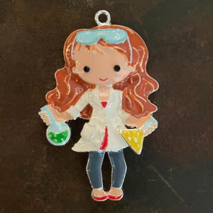 May include: A colorful enamel charm featuring a cartoon scientist with auburn hair, wearing a white lab coat, blue safety glasses, and holding a flask and a beaker. The charm has a silver-colored loop at the top for hanging.