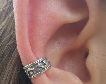May include: Silver ear cuff with a detailed design, worn on the ear. The cuff features a pattern of crescent moons and stars. The jewelry is worn on the upper ear cartilage.