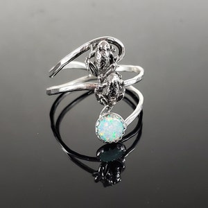 May include: A silver ring with two frog figurines and a white opal gemstone. The ring is designed with a twisted band.