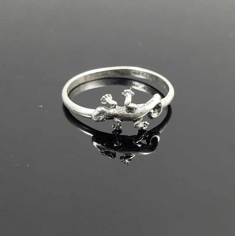 May include: A silver ring with a detailed lizard design. The lizard is facing to the right and has its tail curled around the ring band.