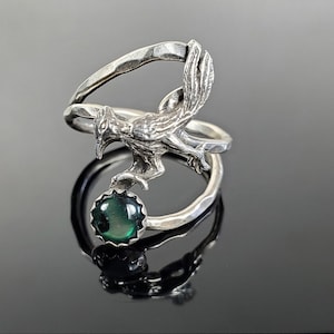 May include: A silver ring with a green gemstone and a detailed bird design. The bird is perched on the ring band, with its wings spread out.