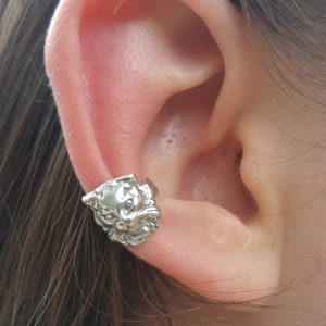 May include: A silver cat head ear cuff earring. The cat's face is detailed with a small nose, eyes, and whiskers.
