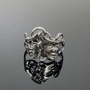 May include: Silver ring with three faces sculpted in relief. The faces are stylized and appear to be wearing masks.