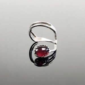 May include: A silver ring with a dark red gemstone set in a silver bezel. The ring is designed with a twisted band.