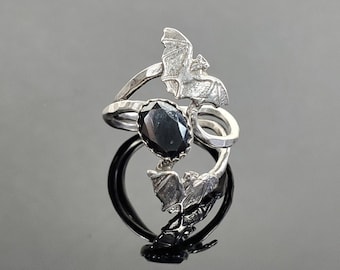 May include: A silver ring featuring a large, oval black gemstone. The ring is adorned with three bat silhouettes, with one bat positioned above the gemstone and two below. The ring has a unique, gothic design.