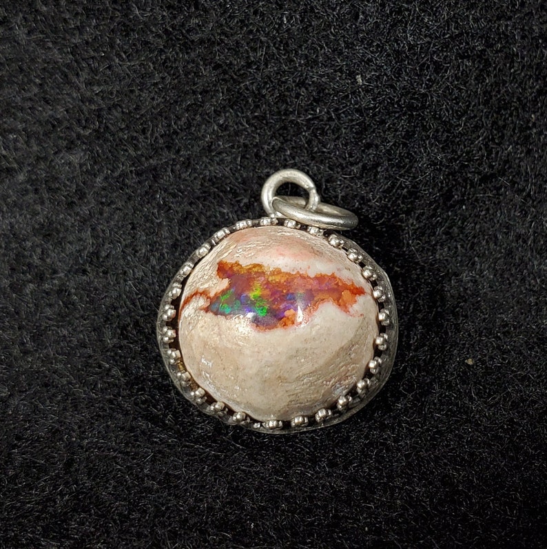 May include: A silver pendant with a round, white opal stone with iridescent flashes of red, green, and blue. The opal is set in a silver bezel with a beaded edge.