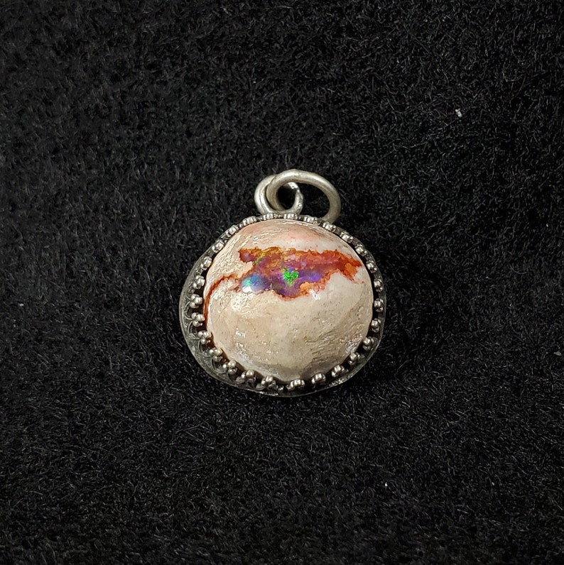 May include: A silver pendant with a round opal stone set in a beaded bezel. The opal has a white base with iridescent flashes of pink, green, and blue.