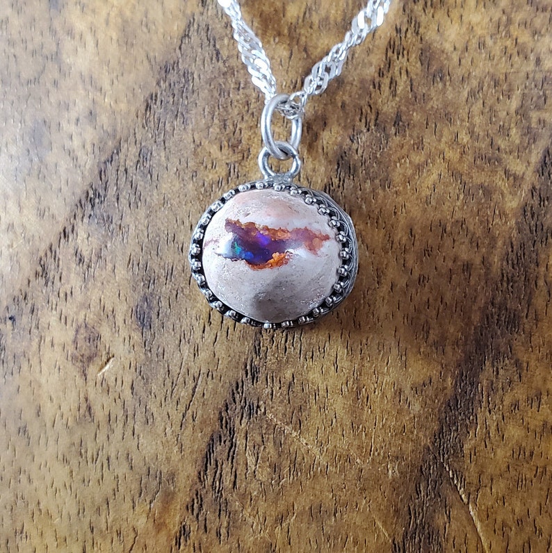 May include: A silver chain necklace with a round pendant featuring a pink and white opal with iridescent blue and purple flashes. The pendant is set in a silver bezel with a beaded edge.