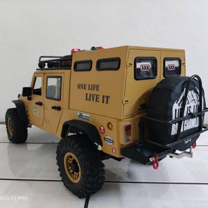 May include: A tan-coloured remote control off-road vehicle with black tyres and a black spare tyre carrier. The vehicle has the text "ONE LIFE LIVE IT" on the side and "RO CRAWL" on the spare tyre cover. It has a roof rack and various stickers.