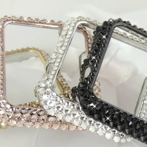 May include: Four bedazzled Apple Watch cases in gold, rose gold, silver, and black. Each case is encrusted with small, sparkling crystals around the square frame. The cases are displayed on a white surface.