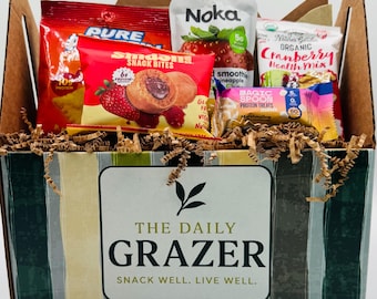 May include: A gift basket filled with various snacks, including chips, fruit snacks, and protein bars. The basket is decorated with a striped pattern and features the text "THE DAILY GRAZER" and "SNACK WELL. LIVE WELL."