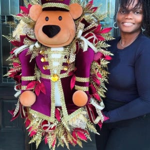 Op de afbeelding: Een decoratieve krans met een pluche teddybeer verkleed als notenkraker in een donkerpaars fluwelen uniform met gouden biezen en knopen. De beer draagt een bijpassende paarse hoed, omringd door gouden varenbladeren en rode lintaccenten.