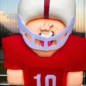 May include: A football player figurine wearing a white helmet with a red center stripe and a light purple face mask. The player has a tan face, a wide open mouth with visible teeth, and a red jersey featuring the white number 10 on the front.
