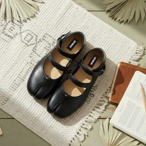May include: A pair of black leather Mary Jane style shoes with a split toe design, resting on a cream-colored rug. The shoes have two straps and the brand name "SOMAE" is visible. The scene includes a cup of coffee, a notebook, and a pencil.
