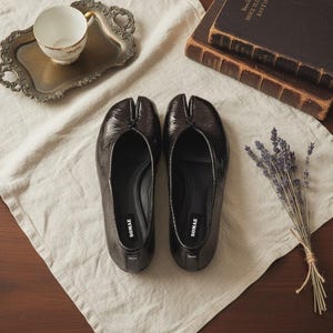 Può includere: Un paio di ballerine lucide marrone scuro con punta divisa e il marchio "SOMAE" sulla soletta. Le scarpe sono esposte su un panno di lino color crema, insieme a un vassoio d'argento vintage con una tazza da tè, vecchi libri e un mazzetto di lavanda.