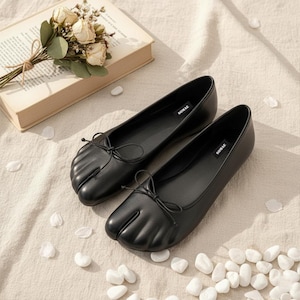 May include: A pair of black leather ballet flats with a split-toe design and small bow detail. The flats are displayed on a neutral surface with scattered white petals and a book with flowers.