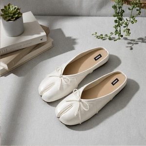 May include: A pair of off-white slip-on shoes featuring a split-toe design, a small decorative bow on the front, and a black label with the text SOMAE printed in white on the inner sole. The shoes have a smooth finish and a low-profile silhouette.