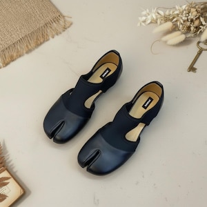 May include: A pair of black split-toe flats with a unique design. The shoes feature a rounded toe box with a split between the big toe and the other toes. The uppers are a combination of smooth and stretchy materials.