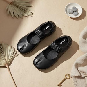 May include: Two black leather ballet flats with a split toe design. The flats feature a ruched vamp and the brand name "SOMAE" is visible on the insole. The shoes are presented on a neutral surface with decorative elements.
