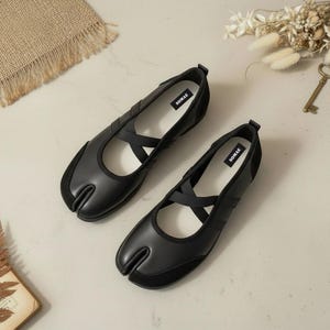 May include: A pair of black split-toe ballet flats with a crisscross strap design. The shoes have a rounded toe and a suede accent on the toe box. The brand name "SOMAE" is visible on the insole.