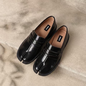 Può includere: Un paio di mocassini neri in pelle con punta divisa. Le scarpe hanno una finitura lucida e una suola spessa. L'interno di ogni scarpa ha un'etichetta con la scritta "SOMAE". Le scarpe sono esposte su una superficie dai toni neutri.