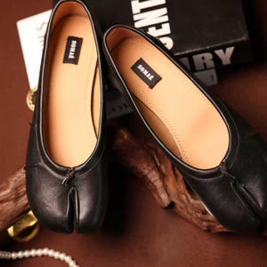 Può includere: Un paio di ballerine in pelle nera con design a punta divisa. Le scarpe hanno una soletta marrone chiaro e una piccola etichetta con la scritta "SONAE". Le scarpe sono esposte su una superficie marrone.