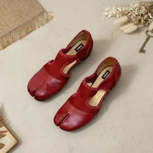 May include: A pair of red leather Tabi shoes with a split toe design. The shoes have a low heel and a cutout detail on the sides. The brand name "SOMAE" is visible on the insole.