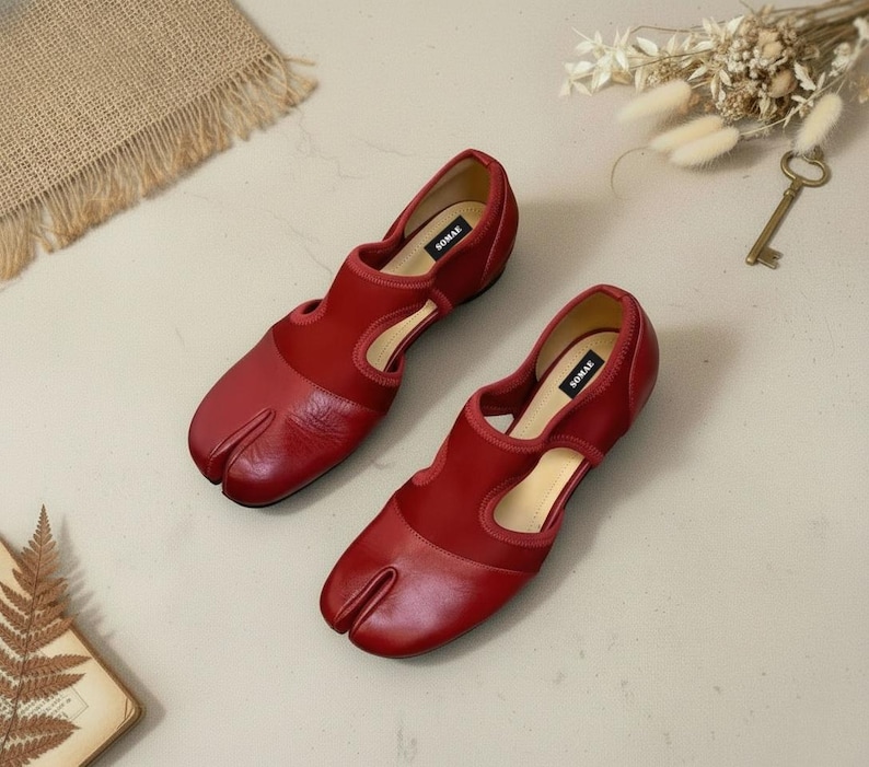 May include: A pair of red leather Tabi shoes with a split toe design. The shoes have a low heel and a cutout detail on the sides. The brand name "SOMAE" is visible on the insole.