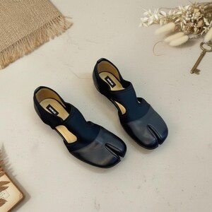 May include: A pair of black split-toe shoes with a unique design. The shoes feature a dark blue fabric upper and a leather toe section. The brand name "SOMAE" is visible on the insole. The shoes are displayed on a light-colored surface.