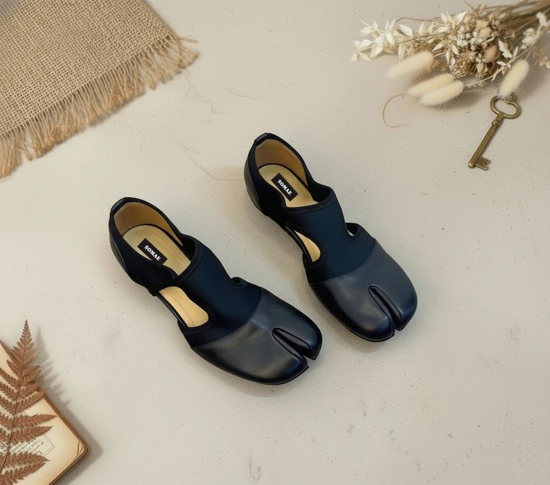 May include: A pair of black split-toe shoes with a unique design. The shoes feature a dark blue fabric upper and a leather toe section. The brand name "SOMAE" is visible on the insole. The shoes are displayed on a light-colored surface.