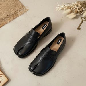 Pode incluir: Um par de mocassins pretos de couro com biqueira dividida. Os sapatos têm um design único com um compartimento separado para o dedão do pé. As palmilhas são bege e a marca "SOMAE" está impressa nelas. Os sapatos estão em uma superfície clara.
