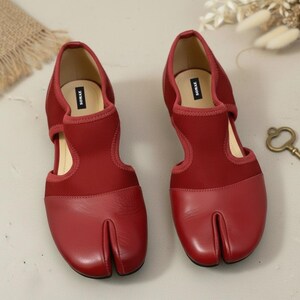 May include: A pair of red split-toe shoes with a unique design. The shoes feature a split toe, a cutout on the sides, and a strap across the top. The shoes are made of a combination of leather and fabric.