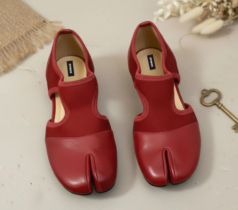 May include: A pair of red split-toe shoes with a unique design. The shoes feature a split toe, a cutout on the sides, and a strap across the top. The shoes are made of a combination of leather and fabric.