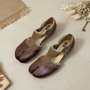 May include: A pair of brown and tan split-toe shoes. The shoes have a unique design with a split between the big toe and the other toes. The shoes have a low heel and a cutout design on the sides. The brand name "SOMAK" is visible on the inner sole.
