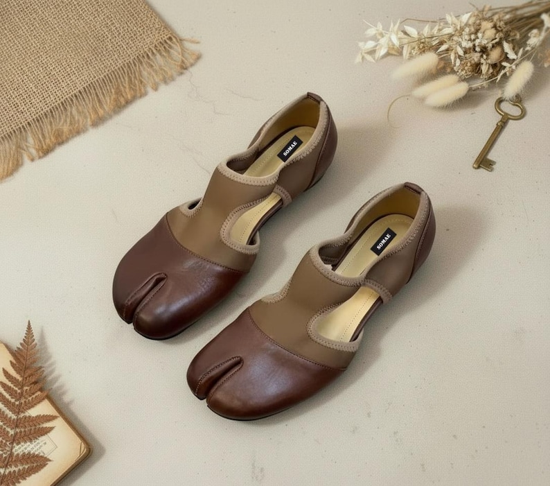 May include: A pair of brown and tan split-toe shoes. The shoes have a unique design with a split between the big toe and the other toes. The shoes have a low heel and a cutout design on the sides. The brand name "SOMAK" is visible on the inner sole.