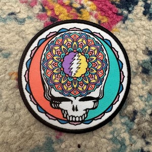Puede incluir: Parche redondo con un cráneo y un diseño de mandala colorido. El cráneo es blanco con detalles negros, y el mandala es una vibrante mezcla de naranja, amarillo, morado y turquesa. El parche tiene un borde negro y un fondo blanco.