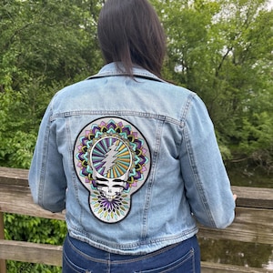 May include: Light blue denim jacket with a large, colorful patch on the back. The patch features a skull design with a lightning bolt and floral mandala elements in purple, green, and yellow. The jacket is worn with dark blue jeans.