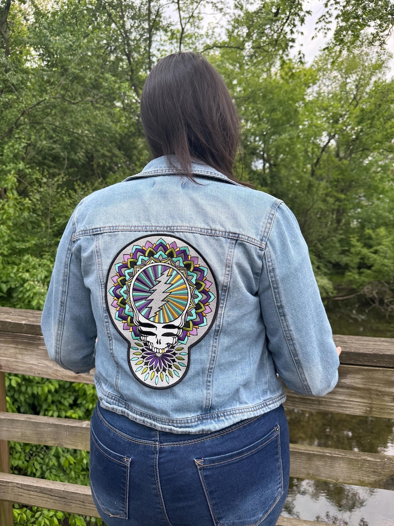 Pode incluir: Jaqueta jeans azul clara com um grande patch colorido nas costas. O patch apresenta um design de caveira com um raio e elementos florais de mandala em roxo, verde e amarelo. A jaqueta &eacute; usada com jeans azul escuro.