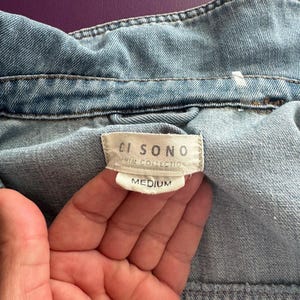 Pode incluir: Close-up de uma jaqueta jeans azul claro. A gola e as costuras da jaqueta s&atilde;o vis&iacute;veis, com uma etiqueta que diz "CI SONO DENIM COLLECTION MEDIUM". O tecido tem uma apar&ecirc;ncia texturizada.