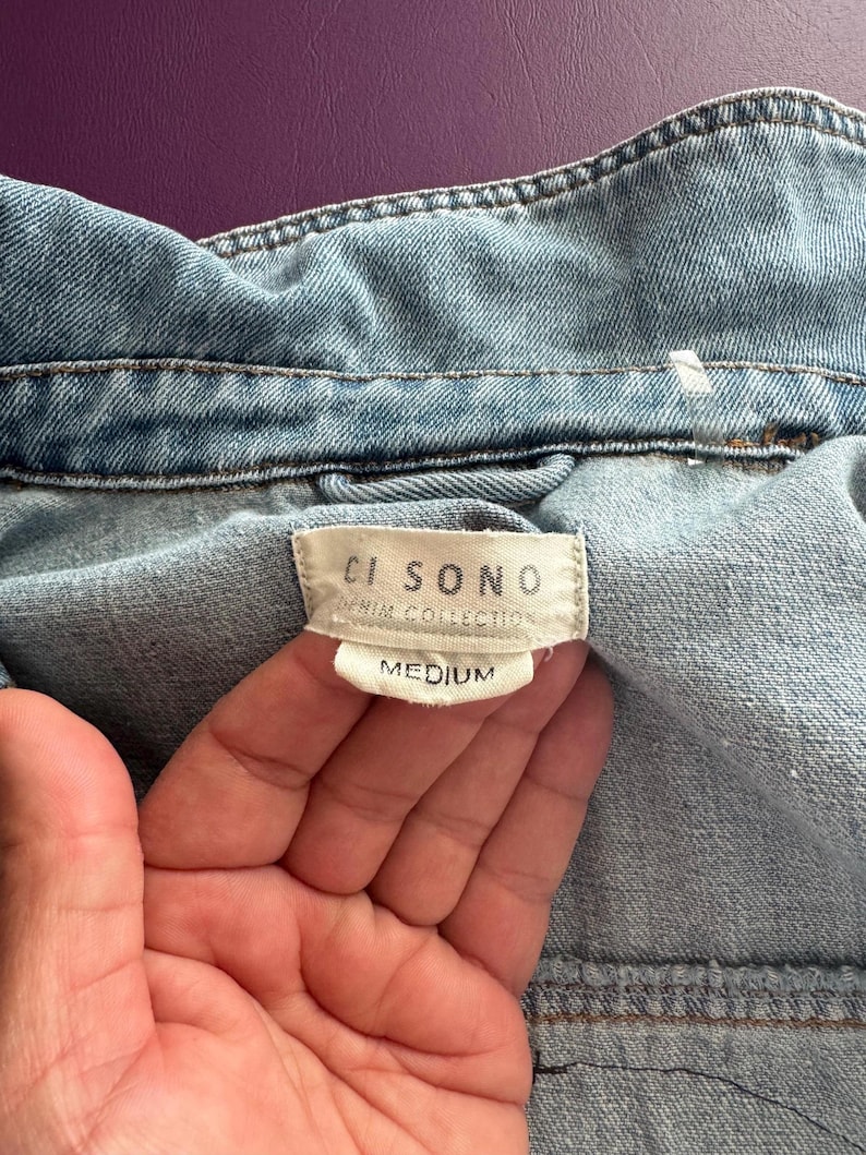 Pode incluir: Close-up de uma jaqueta jeans azul claro. A gola e as costuras da jaqueta s&atilde;o vis&iacute;veis, com uma etiqueta que diz "CI SONO DENIM COLLECTION MEDIUM". O tecido tem uma apar&ecirc;ncia texturizada.