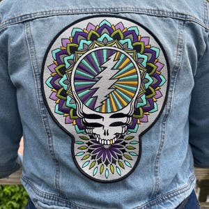 Pode incluir: Casaco de ganga azul claro com um grande patch colorido nas costas. O patch apresenta uma caveira estilizada com um raio e um design de mandala em tons de roxo, verde-azulado, amarelo e verde.